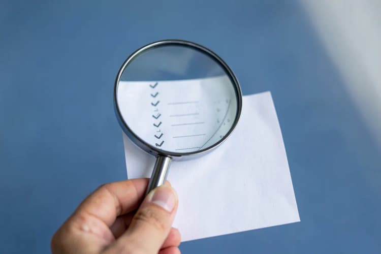 Mão segurando uma lupa sobre uma lista de opções com checkmarks, simbolizando a escolha criteriosa de uma agência.