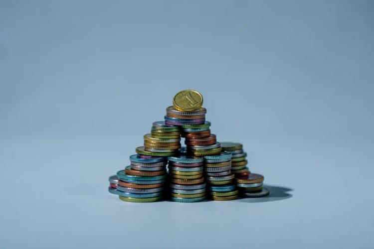 Pilhas de moedas de diferentes tamanhos e cores empilhadas em forma de pirâmide, com uma moeda dourada no topo.