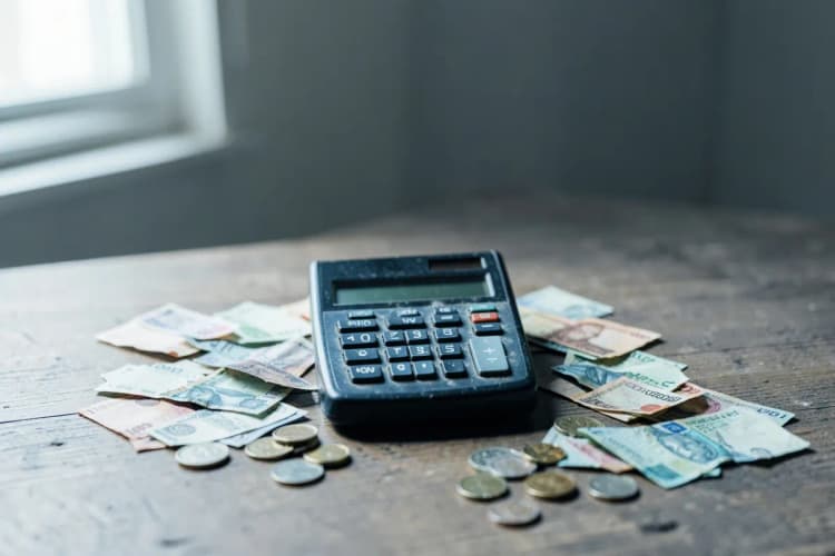 Calculadora antiga sobre uma mesa de madeira com algumas notas de dinheiro e moedas espalhadas, luz de janela lateral.